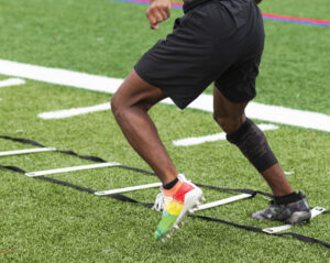A high school boy is running ladder drills with colorful cleats on a turf field during summer camp practices.